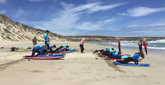 Two groups of students in a row on surfboards on a beach.