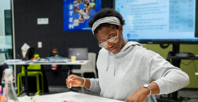 Female student in an indoor workshop setting looking down and arranging unseen items on a table.