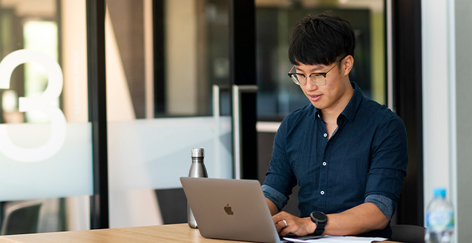 Photo of a student wearing glasses and a button-up shirt as they sit at a desk and use their laptop. There are glass sliding doors in the background.