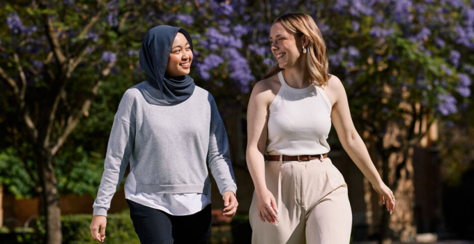 Photo of two female students walking on campus with jacaranda trees in the background.