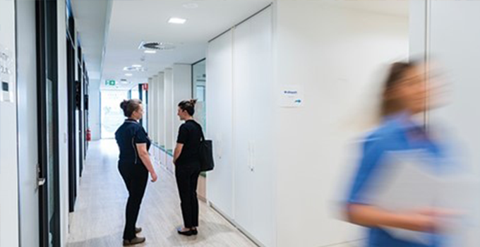 Two female nurses standing and talking in a medical clinic corridor with another nurse walking outside of the frame.