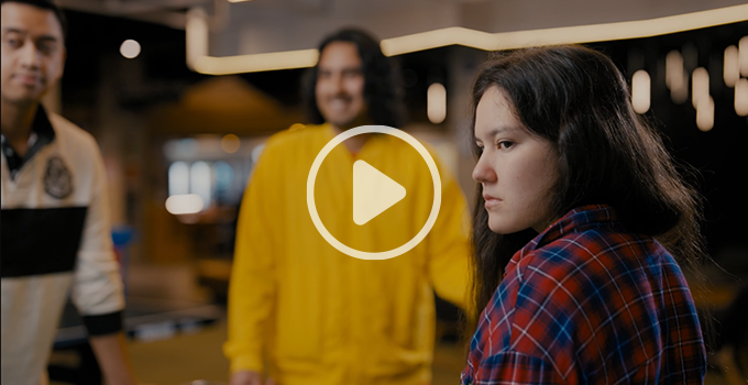 Still frame of Active Bystander video "Step up. Speak Out. – Rape Culture is Dangerous." It features two males and one female student at City West student lounge. The female student in foreground is looking contemplative at something off screen. The males are out of focus in the background, one is looking concerned while the other is laughing. Overlayed over the still is a semitransparent play icon.