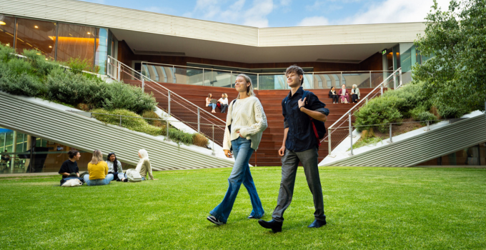 Two smiling university students walking across the lawn infront of Pridham Hall. Groups of students sit on the grass and on the tiered seating in the background.