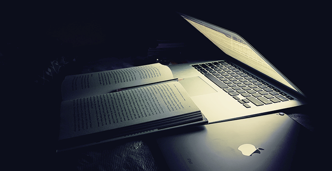 Half open laptop emitting light in a dark room. A open text book and a second closed laptop are illuminated as a result. 