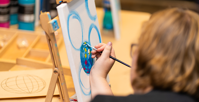 Over the shoulder photo of a student painting on an A4 canvas propped up on a easel.
