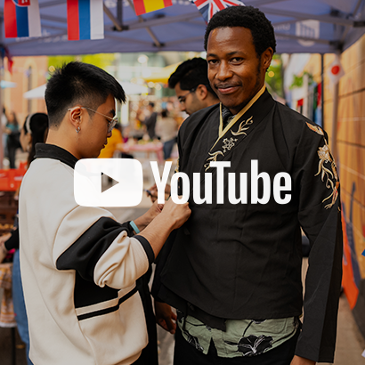 Image of a male student fitting a traditional outfit on another male student at Multicultural Festival, with YouTube logo as an overlay on the image