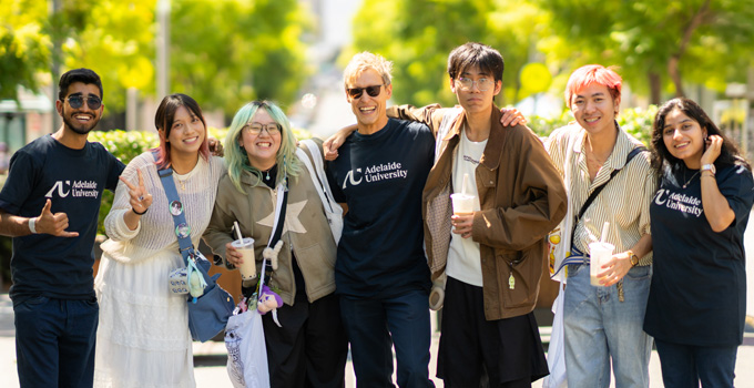 Mixed group of seven students smiling to camera in sunny outdoor setting. A few of them a wearing an Adelaide University branded tee.