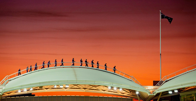Wide angle image of a large group of people walking across Adelaide Oval rooftop  during a stark orange sunset.