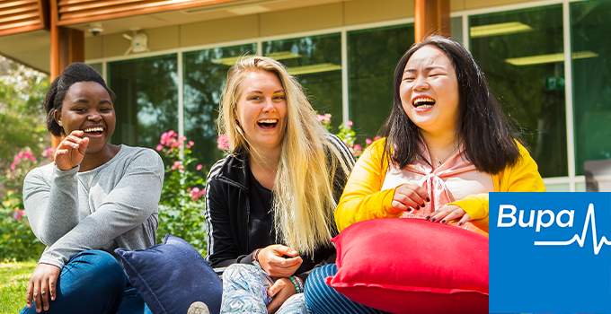 Photo of three students laughing as they sit together on the grass outside a building with floor-to-ceiling windows. The Bupa logo sits in the bottom-right corner.