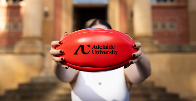 Photo of person holding out a red AFL football over their face and directly to camera. The football is branded with the Adelaide University logo.