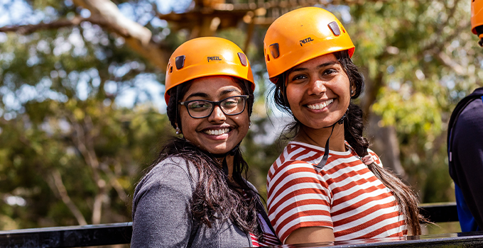 Two female students wearing orange safety helmets, awaiting tree climb 