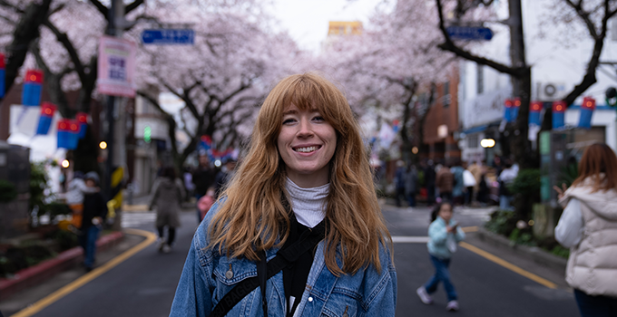 Smiling student standing on a street with cherry blossom trees in the background. People are walking across the road and on the sidewalk.