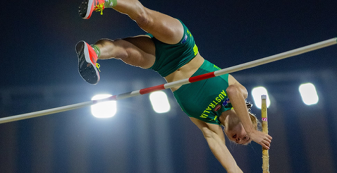 Action shot of pole vaulter mid-air wearing a green and gold Australian uniform.