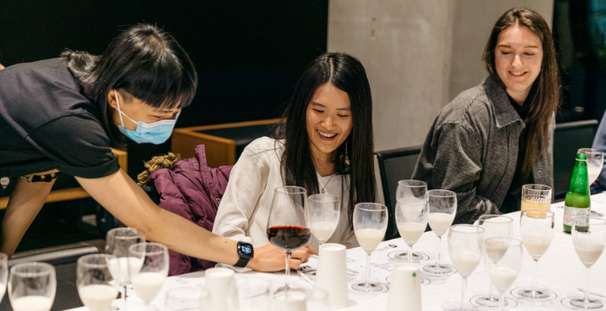 Two people sit at a table, surrounded by wine glasses filled with different plant-based milk. A third person leans over, pointing to something on a page. Everyone is smiling.