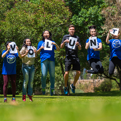Image of male and female students jumping up outdoors holding letters that spell out 'Volunteer'.