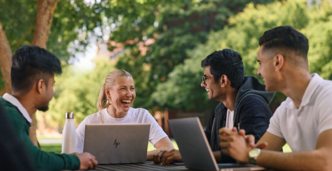 Photo of four students chatting and laughing while sitting around a table with laptops