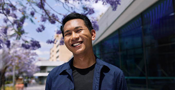 Image of a male student smiling in front of Pridham Hall surrounded by jacaranda flowers