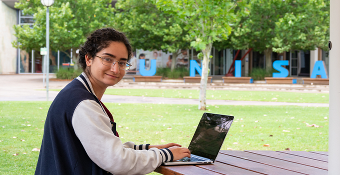 Female student studying on laptop outside in front of UniSA letters at Mawson Lakes Campus