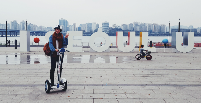 Student on segway scooter posing in front of Seoul letter sign and city skyline.
