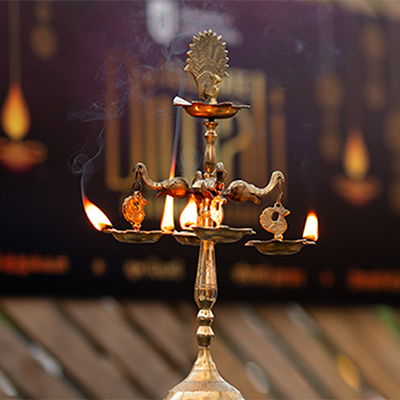 Lit oil lamp with UniSA Diwali branded corflute out of focus in the background.