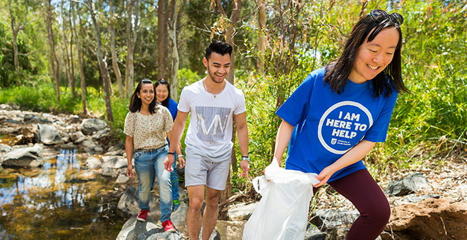 Small group of student volunteers walking along a creek. The student in the foreground is carrying a rubbish bag and wearing a blue UniSA shirt.