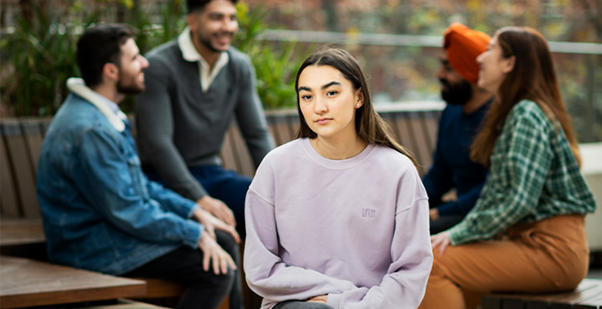 Female student facing the camera and seperate from a group of students chatting in the background. Everyone is sitting at the City East Plaza.