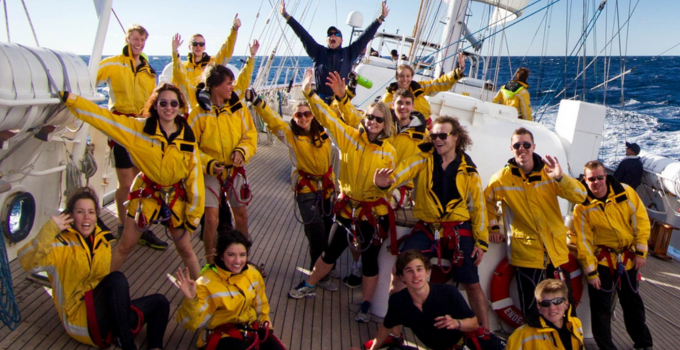 A group of over a dozen students smiling and posing enthusiastically while on a sailing ship out at sea.