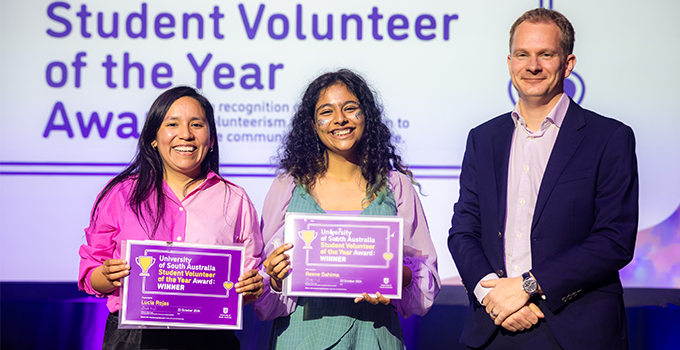 Student Volunteer of the Year co-winners Reene Dahima and Lucia Rojas on stage, holding their certificates to camera. They are joined by Deputy Vice Chancellor Student Experience & Success Tom Steer.