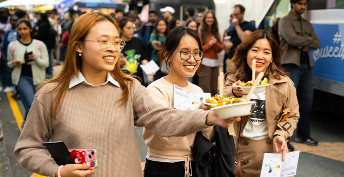 Three female students holding food with an outdoor event crowd in the background.