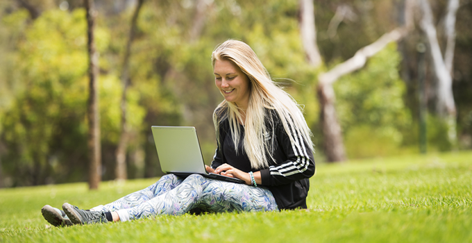 Photo of smiling student using their laptop while sitting outside on the grass.