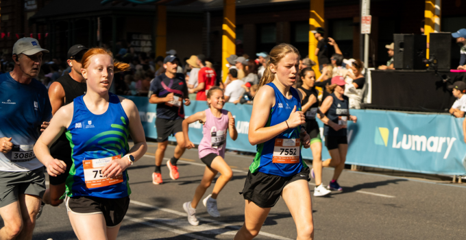 Two UniSA students running in the City to Bay Fun Run. Both runners are wearing UniSA Sport singlets and race number labels, and they are followed by other runners of all ages. A crowd of people  stands behind a Lumary banner in the background.