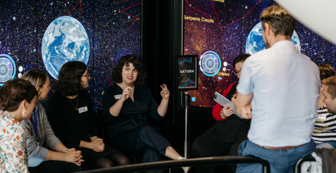 Eight people with name tags sitting in a circle in front of large display screens. The MOD. TV screens show stars, constellations and planets. The group are listening intently to one person who is speaking.