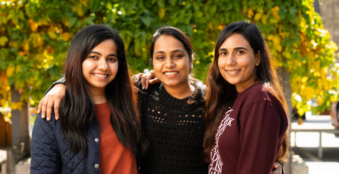Photo of three female students standing close together on campus, smiling at the camera with arms around each other