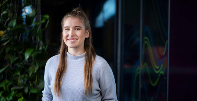 A young woman with long straight hair smiles at the camera while standing indoors