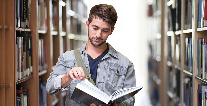Male student standing in library aisle reading a book.
