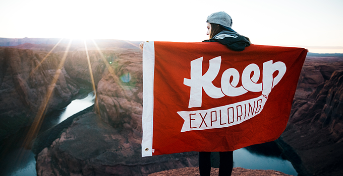 Image of female looking over canyon vista holding a red "Keep Exploring" flag.