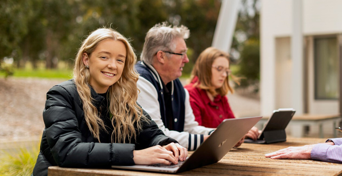 Image of male and female students on their laptop on a table outside their campus building