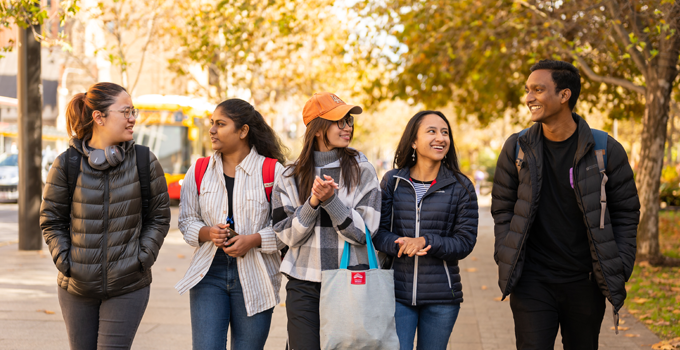 Five international students talking to one another as they walk along a footpath. There are trees and a street with cars and a bus in the background.